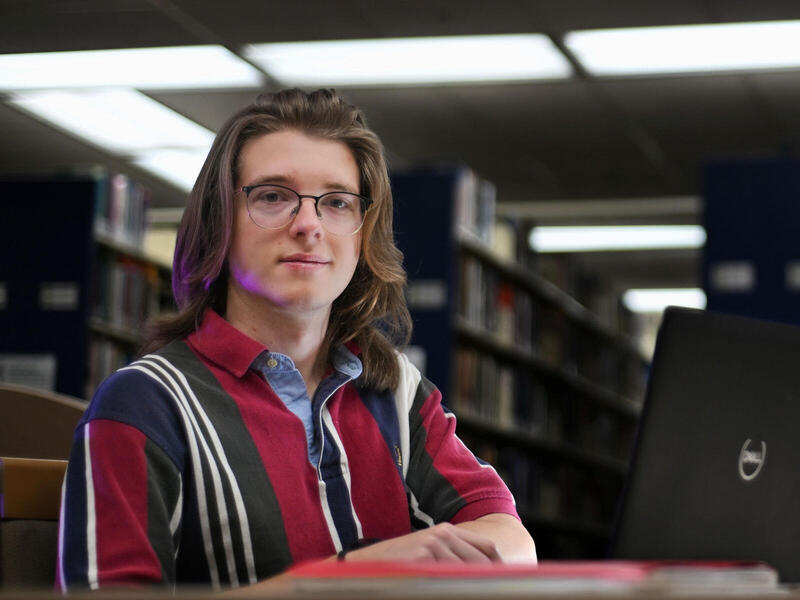 Man in glasses sitting at a table in a library with an open laptop.