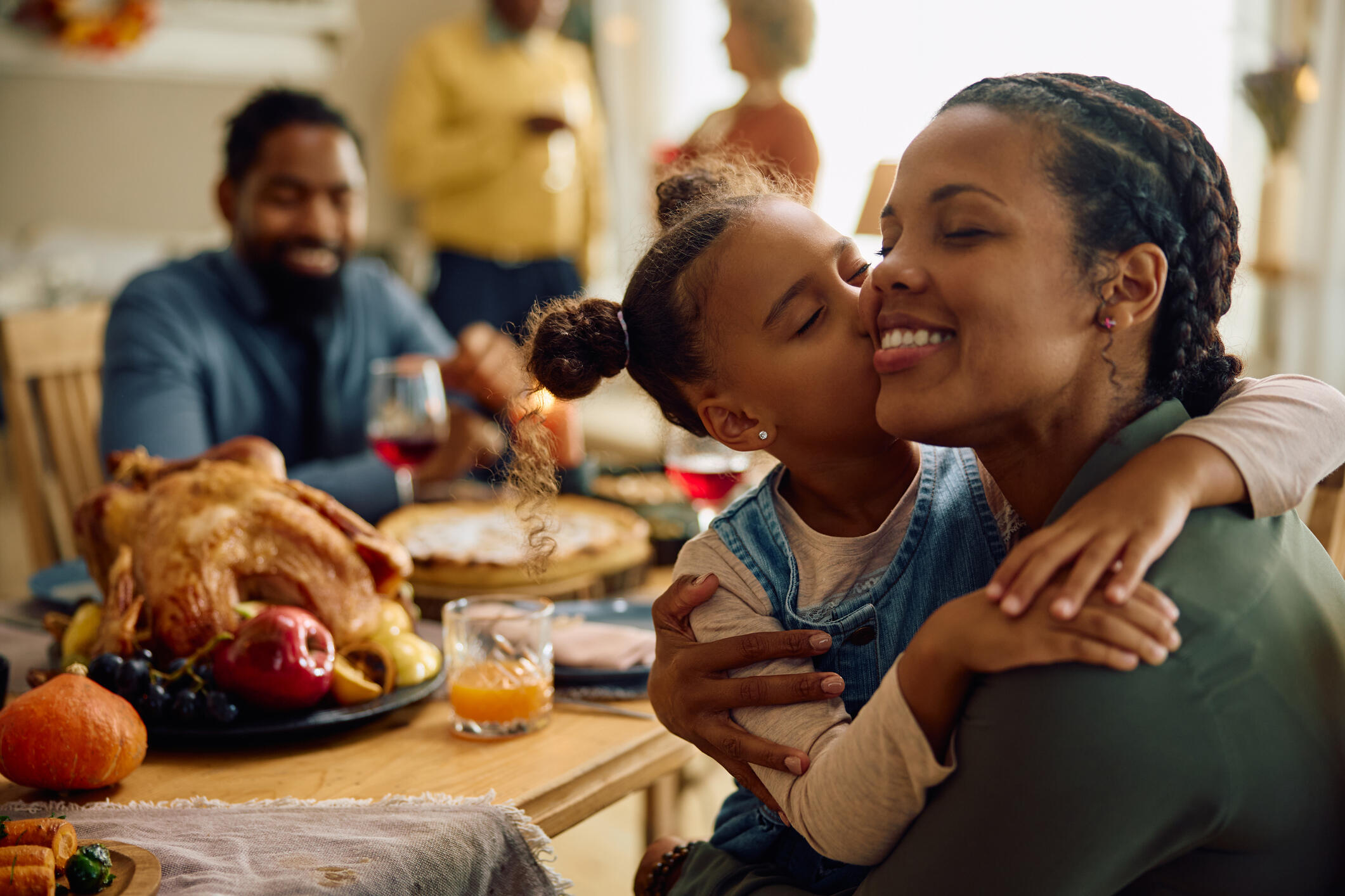 A photo of a family at the Thanksgiving dinner table. A little girl is kissing her mom on the side of her face. A dad is sitting across from them, smiling. 