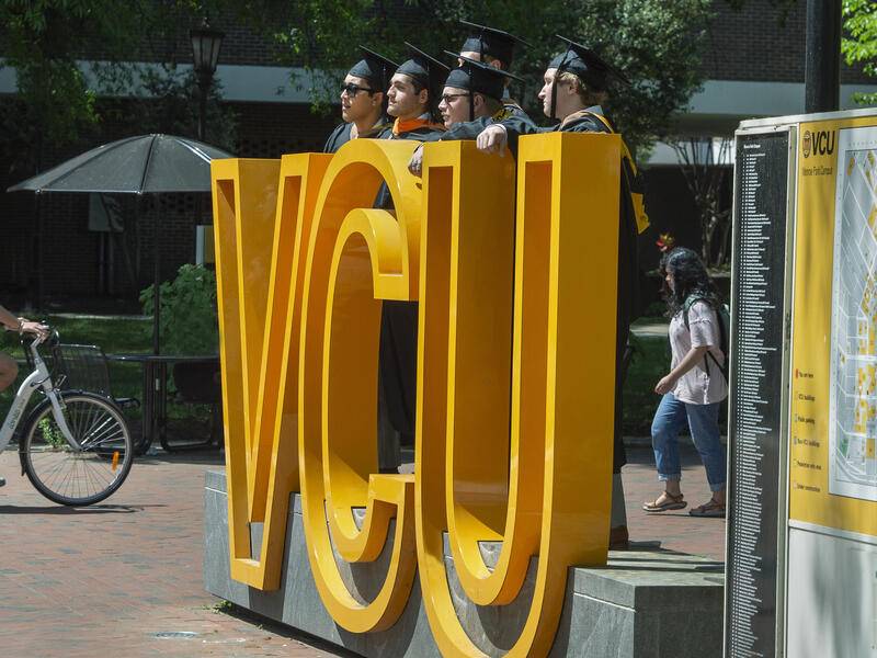 A photo of five people wearing graduation gowns and caps standing behind a sign made up of giant yellow letters that spell out \"VCU.\" To the left of the sign is a man riding his bike past it and to the righ is a woman walking. 