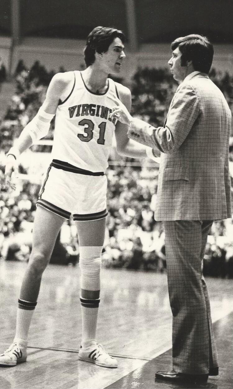 A black and white photo of a basket ball player on a court talking to a man in a suit. 