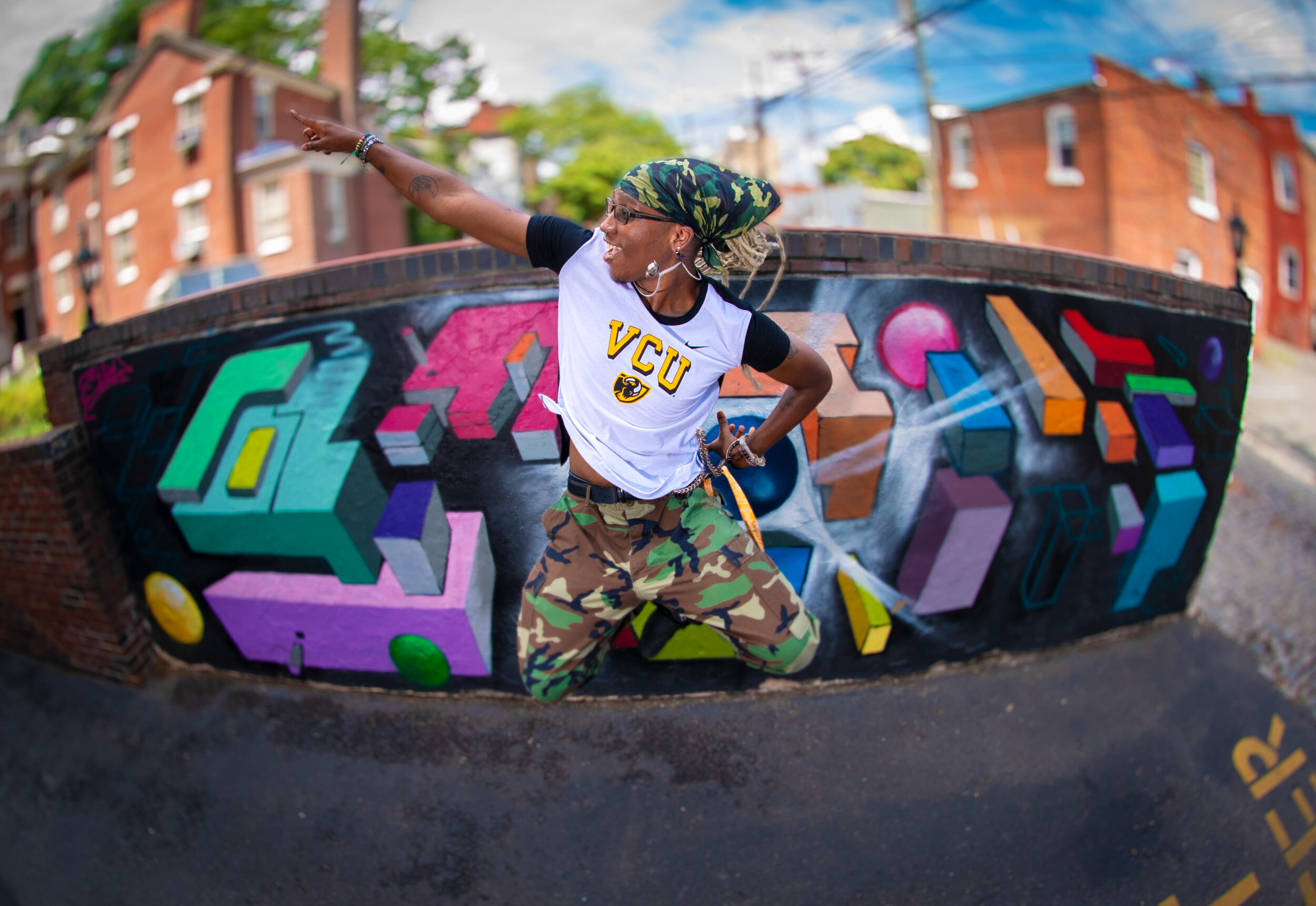 A woman in a VCU shirt and camouflage pants jumps in front of a colorful mural and points upward.