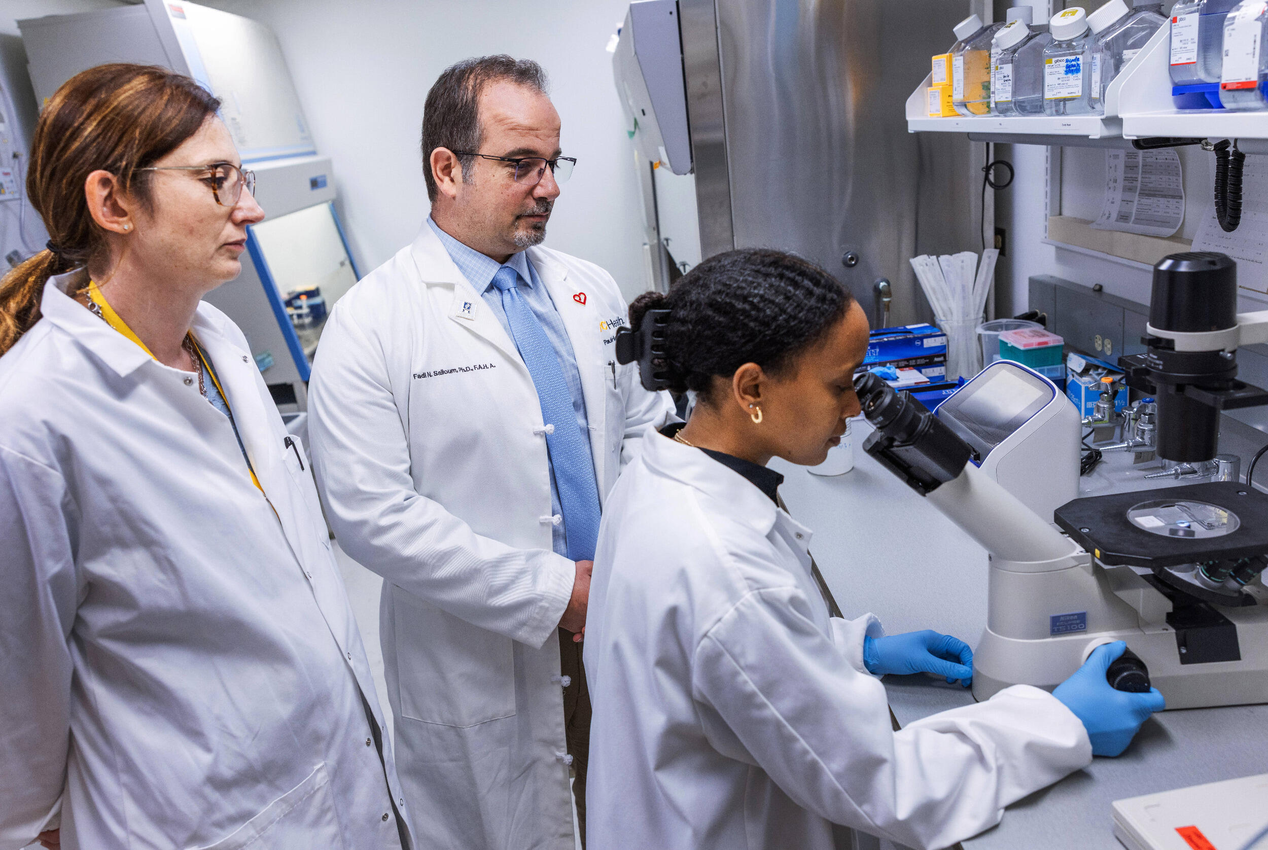 A woman and a man in lab coats stand and observe a woman in a lab coat who is looking through a microscope.