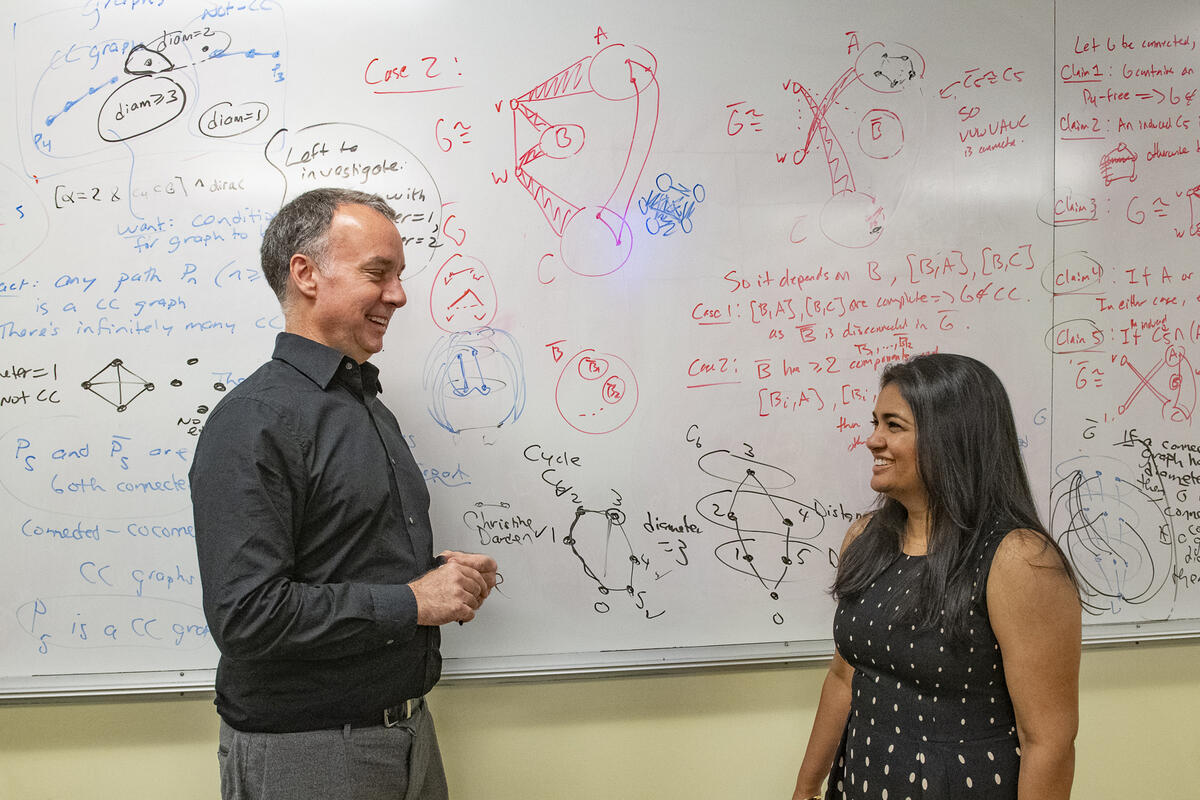 A photo of a man and woman facing each other and smiling. They are standing in front of a white board covered in math equations. 