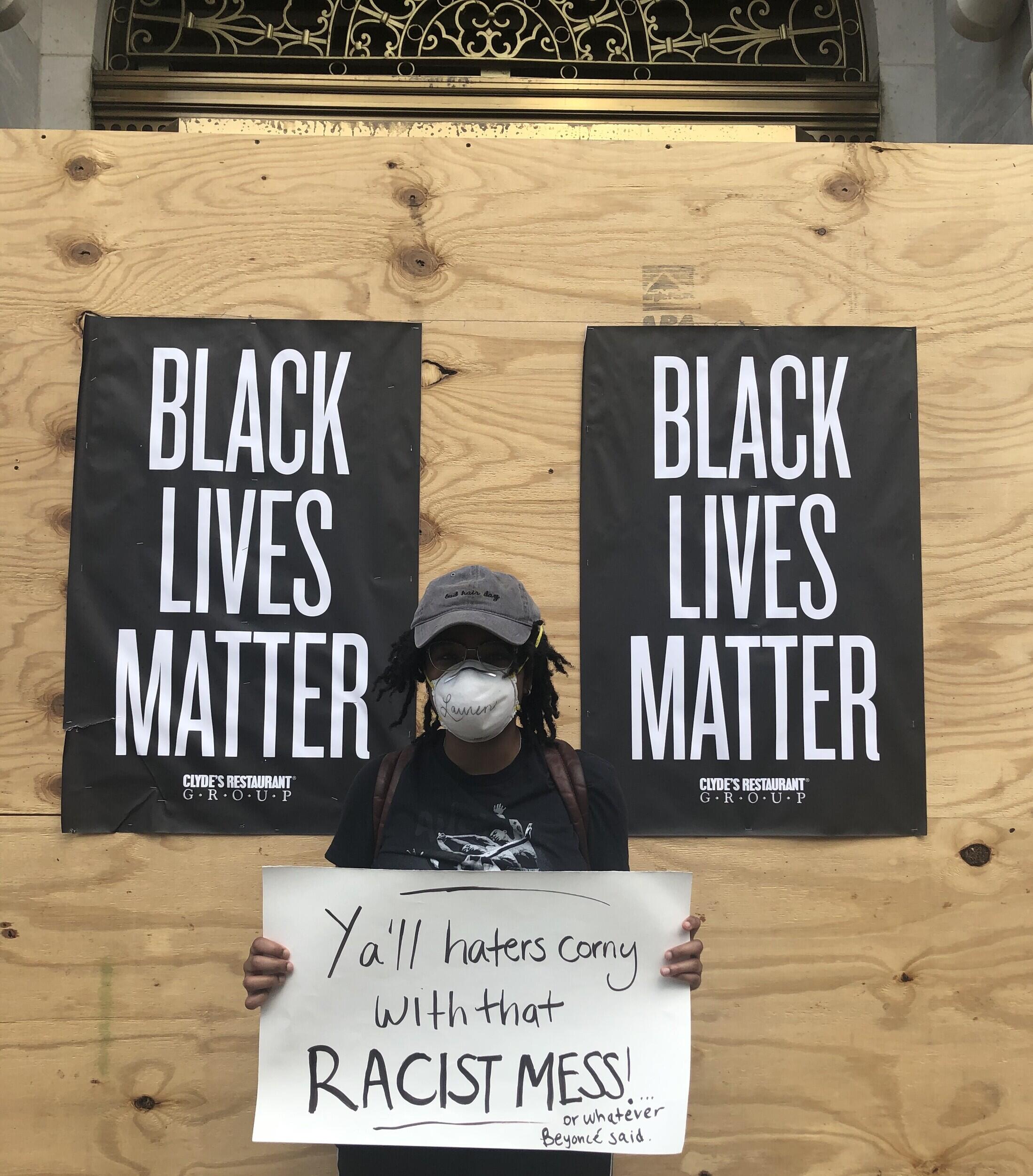 A person holding a sign in front of plywood boards with Black Lives Matter posters. The held sign reads "Y'all haters corny with that RACIST MESS! or whatever Beyonce said."