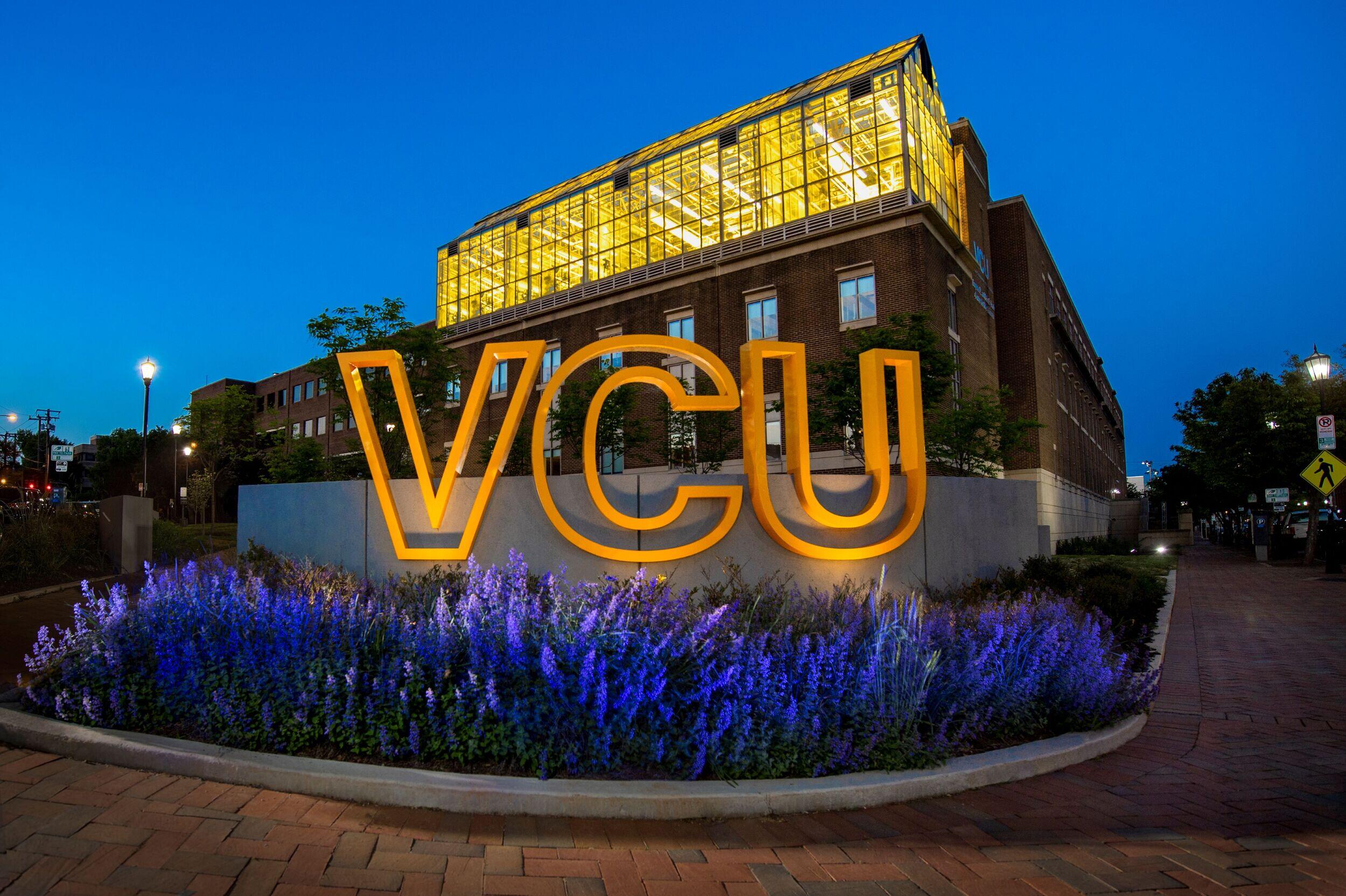 A photo of a sign with giant letters that spell out \"VCU\" in front of a building lit up at dusk. 