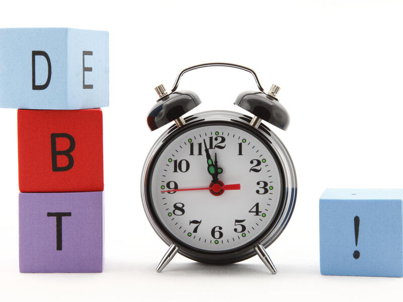 A photo of an alarm clock with multicolored building blocks on either side of it. The three blocks on the left side spell out \"D\" \"B\" \"T\" and the one on the right has an exclamation point on it.