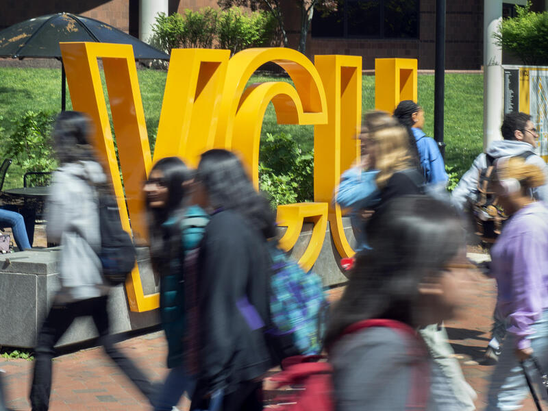 A crowded sidewalk with people walking by. Behind the people is a sign that says \"VCU\" in large yellow letters. 