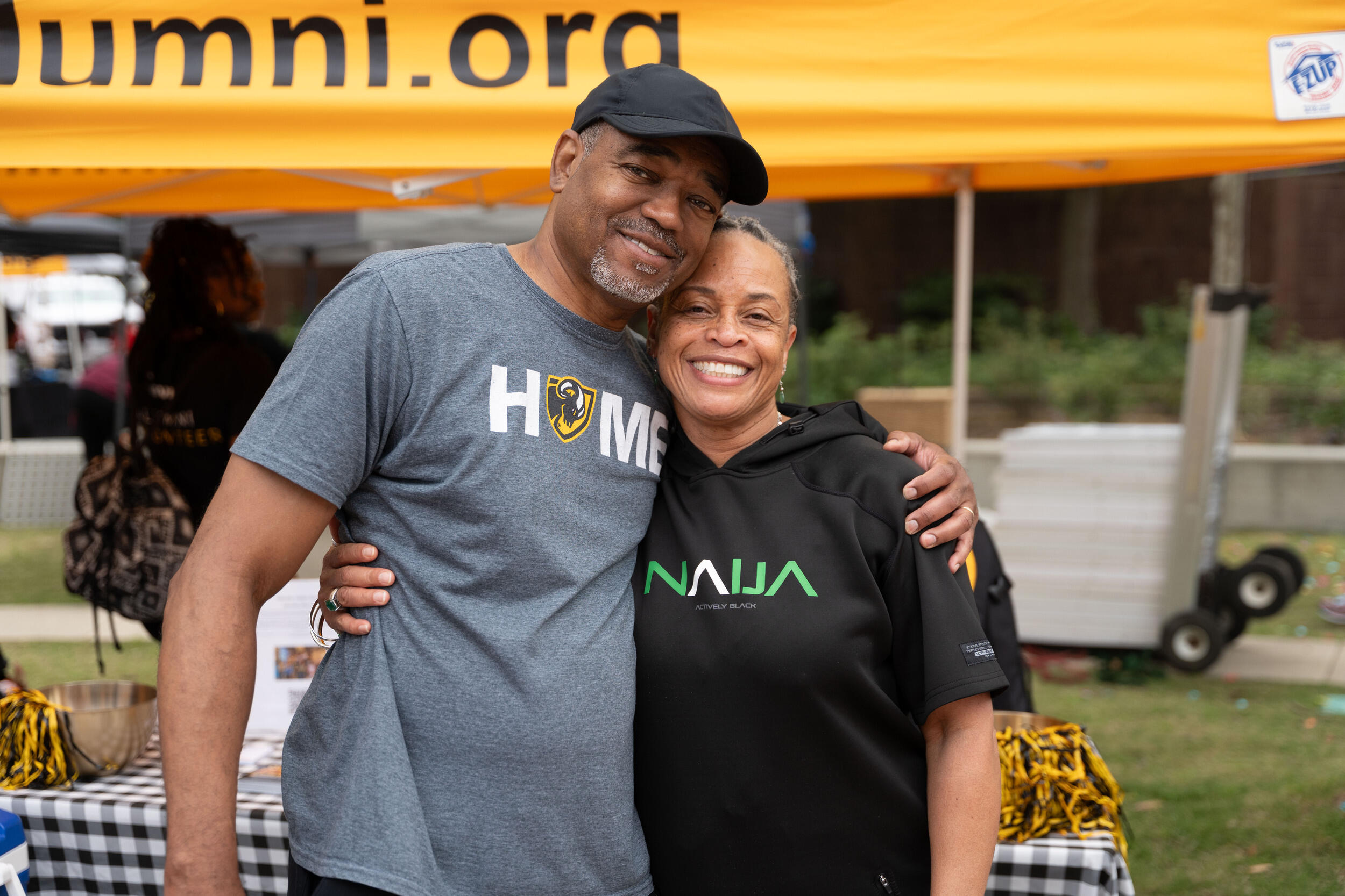 A photo of a man and woman standing next to each other. The man has his arm around the woman's shoulder, and the woman has her arm around his torso. 