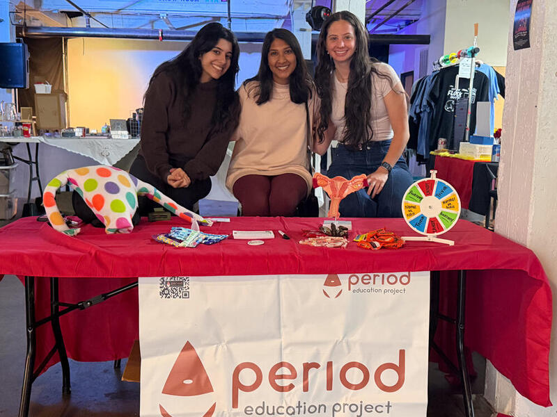 A photo of three women standing behind a table with a red table cloth on it. A banner on the front of it says \"period education project\" with an illustration of a blood drop wearing underwear. 