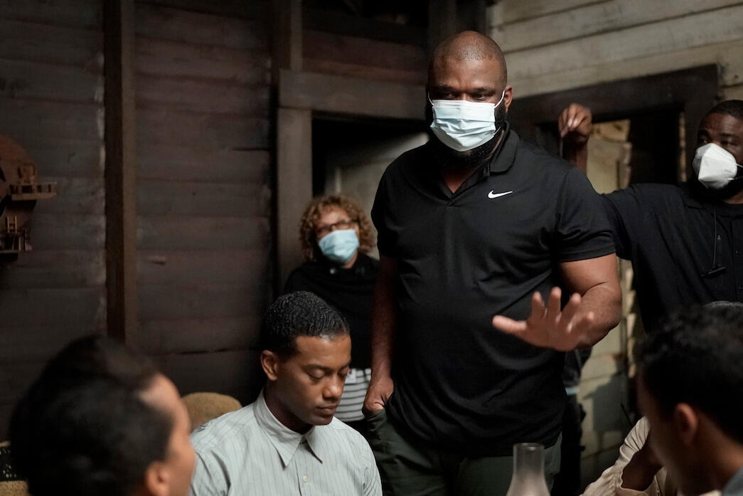 A man in a surgical mask stands and speaks with actors sitting around a table.