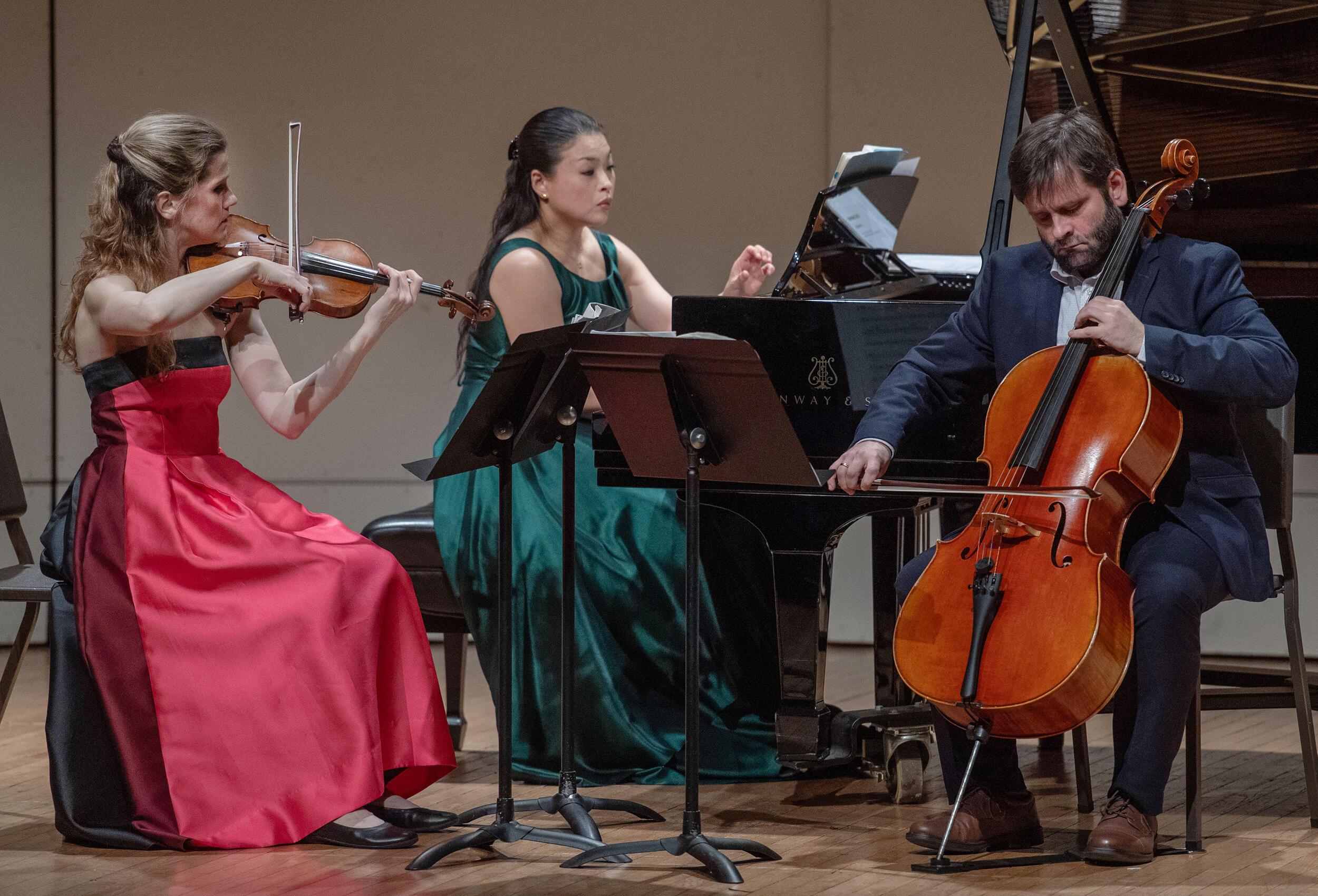 A photo of three people sitting in a semi circle. The people on the left and right are playing string instraments. The person in the middle is playing the piano. 