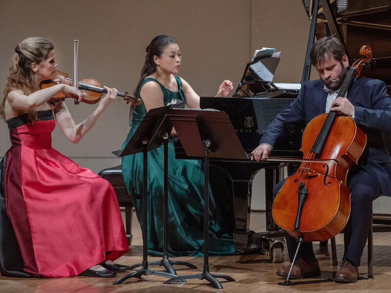 A photo of three people sitting in a semi circle. The people on the left and right are playing string instraments. The person in the middle is playing the piano. 