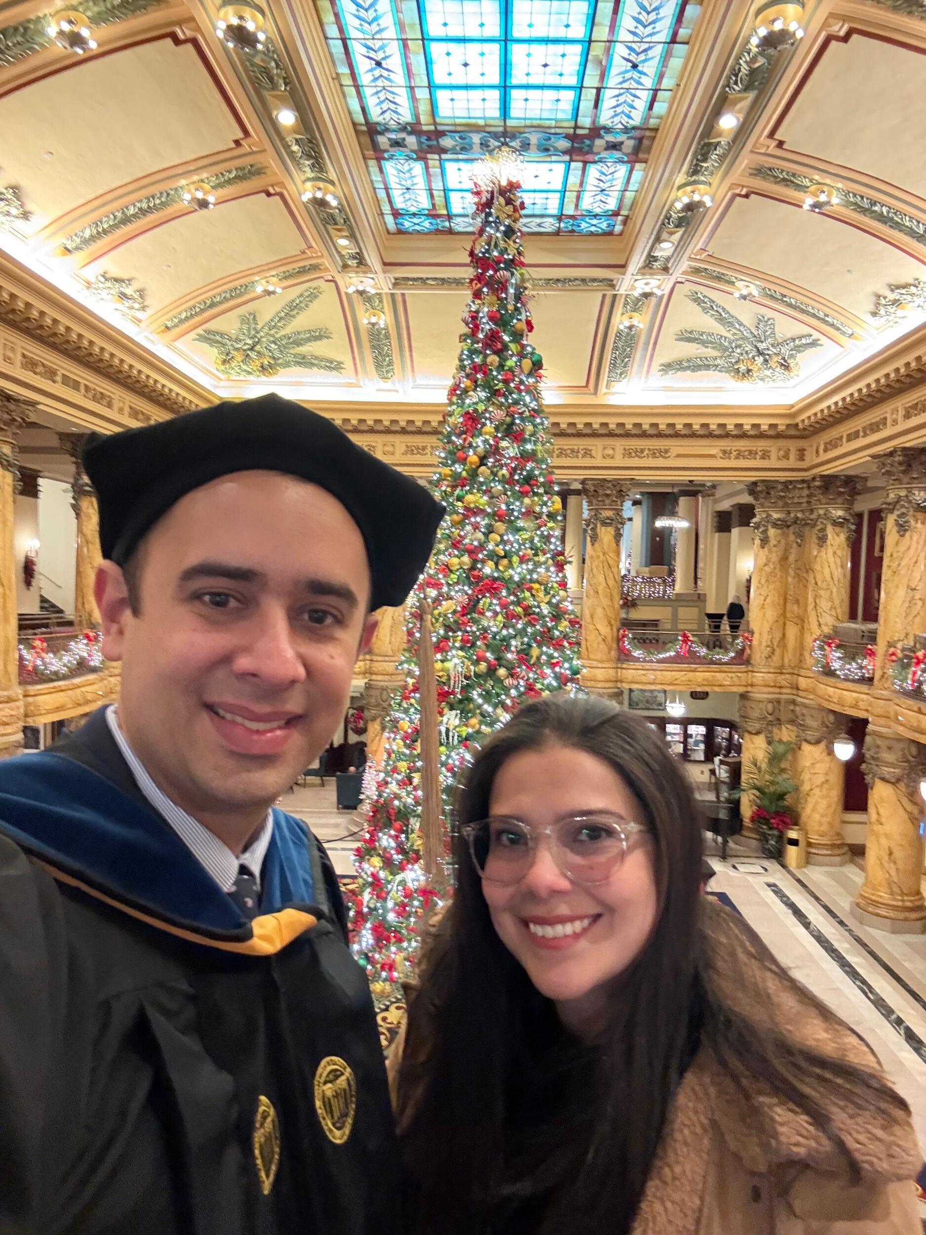 A photo of a man in a graduation cap and gown standing next to a woman in a coat and scarf standing in front of a large Christmas tree. 