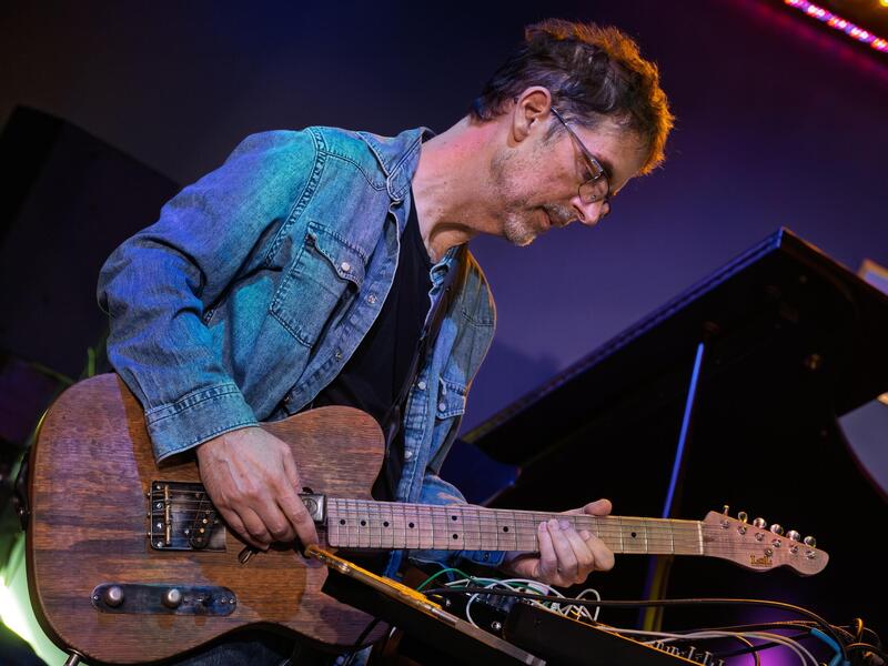 A photo of a man playing a wooden electric base guitar