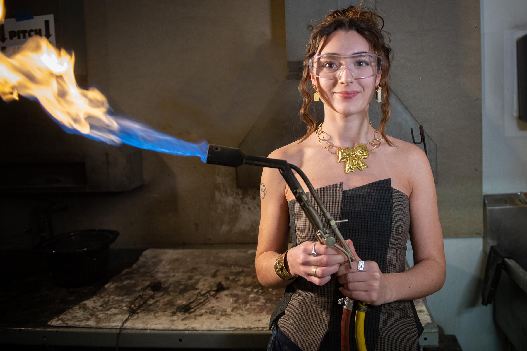 A photo of a woman wearing safety goggles and holding a blow torch while wearing a stylish top, a necklace, and earrings. 