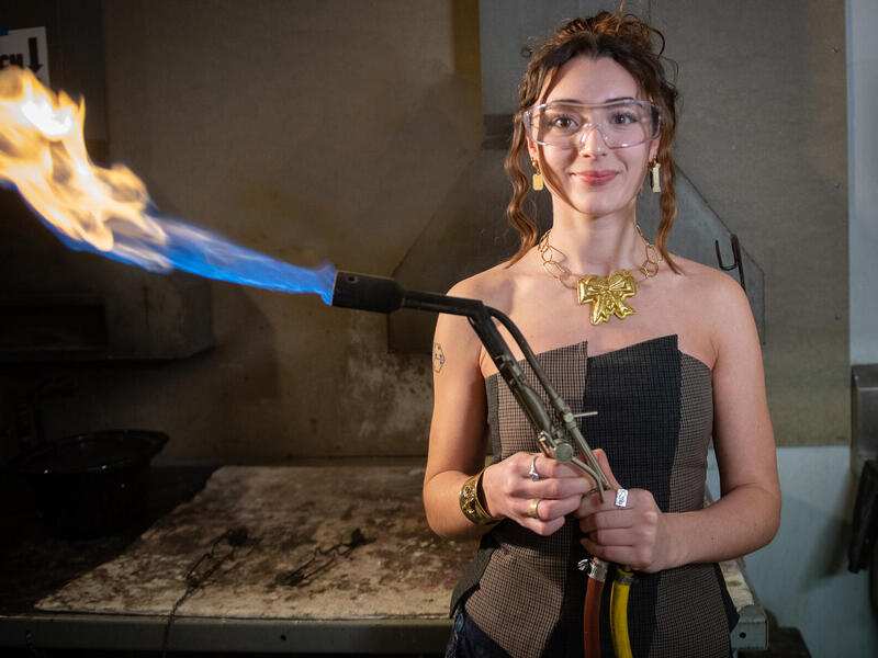 A photo of a woman wearing safety goggles and holding a blow torch while wearing a stylish top, a necklace, and earrings. 