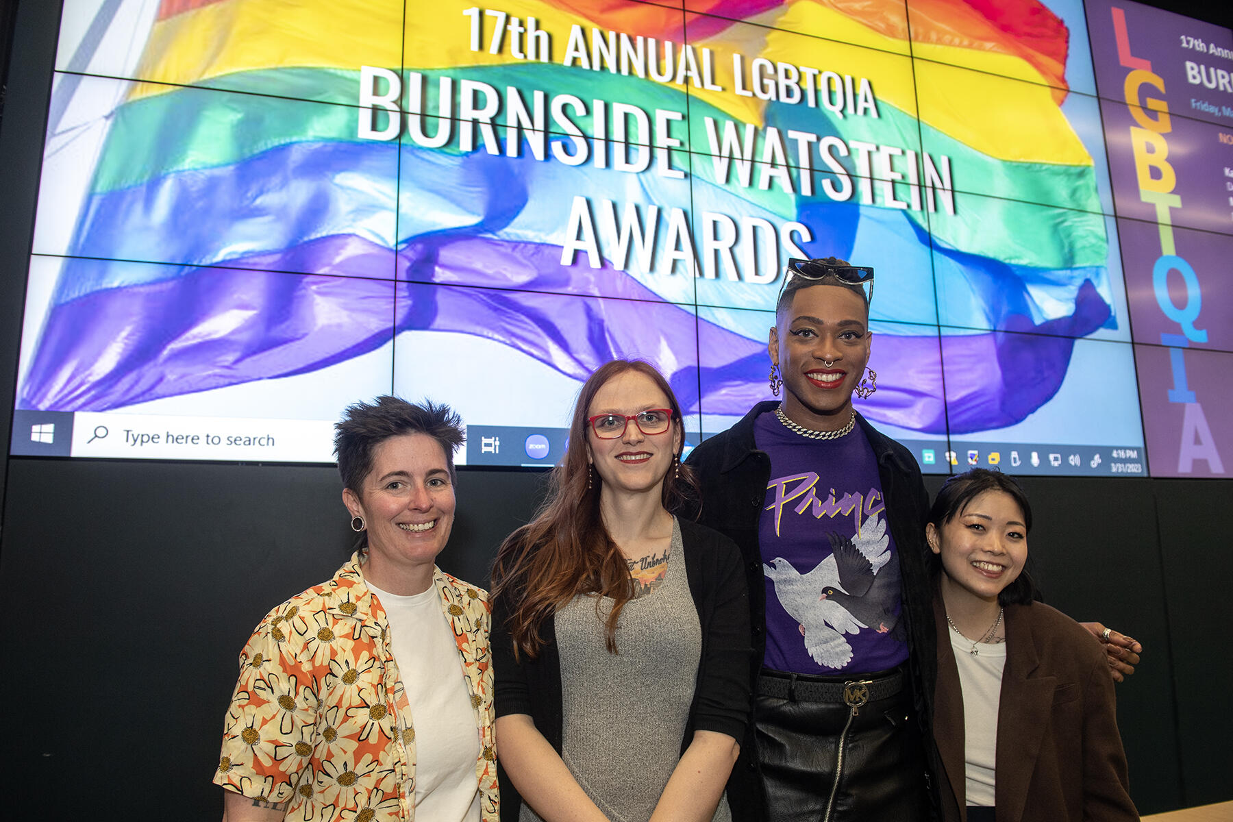 Four people standing in front of a screen with a rainbow flag and white text that reads \"17th ANNUAL LGBTQIA BURNSIDE WATEIN AWARDS\"