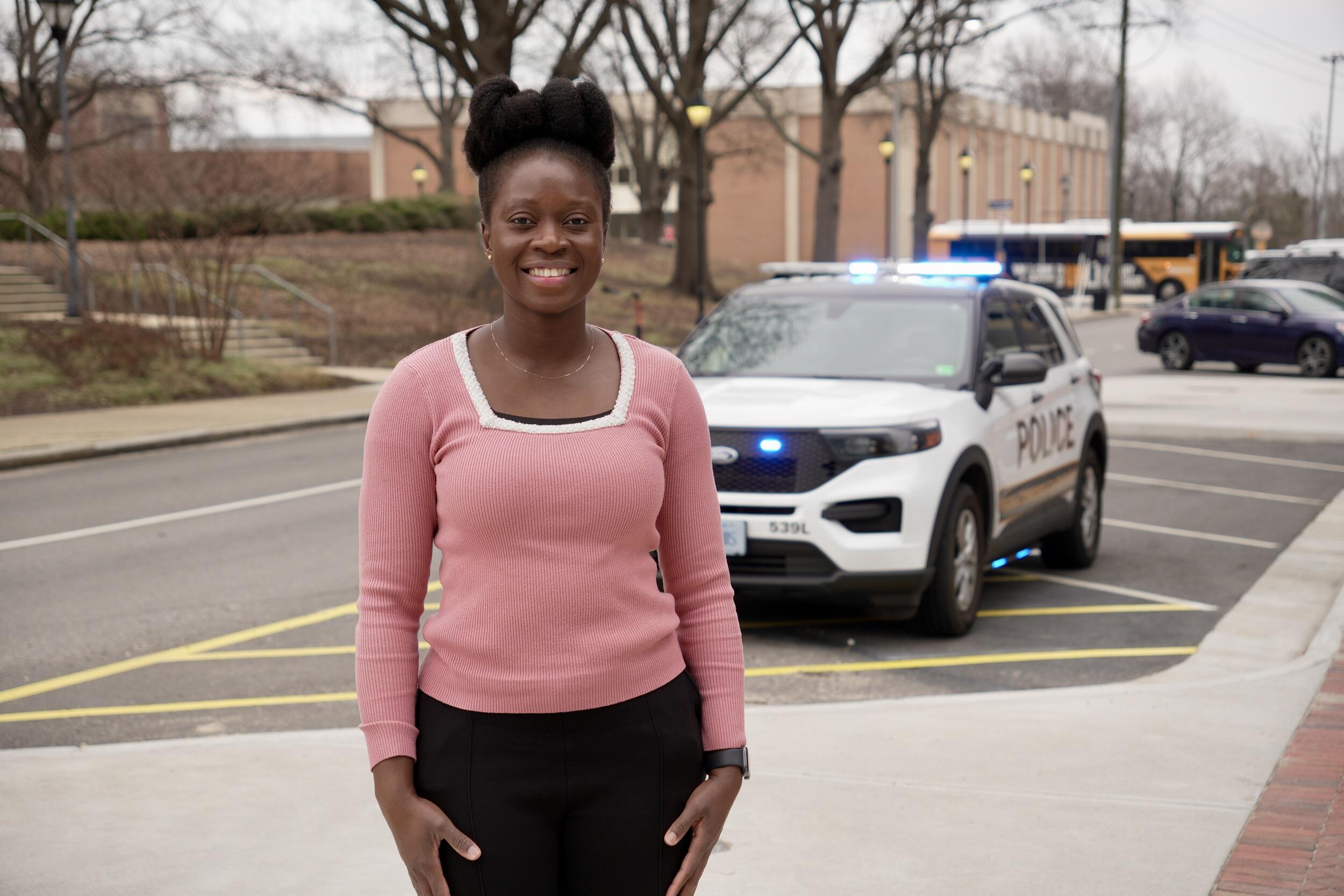 A photo of a woman from the hips up standing and smiling. Behind her is a police SUV> 