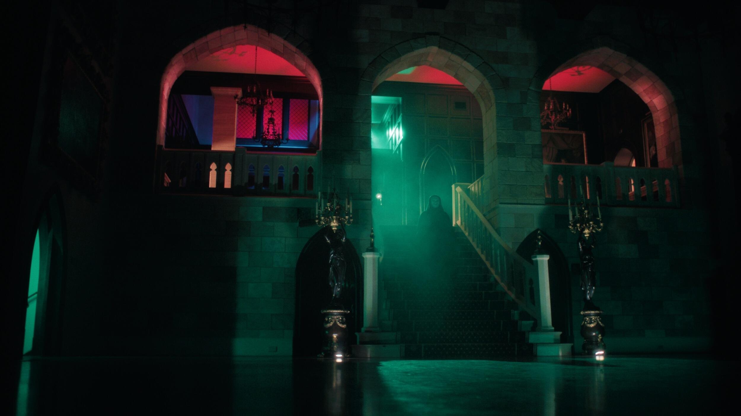 A photo of a hallway that is dark other than green and red light coming through windows. 