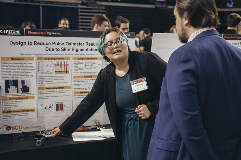 A photo of a woman talking to a man in front of a poster board sitting on a table. She is reaching over to the table and touching a pulse oximeter.