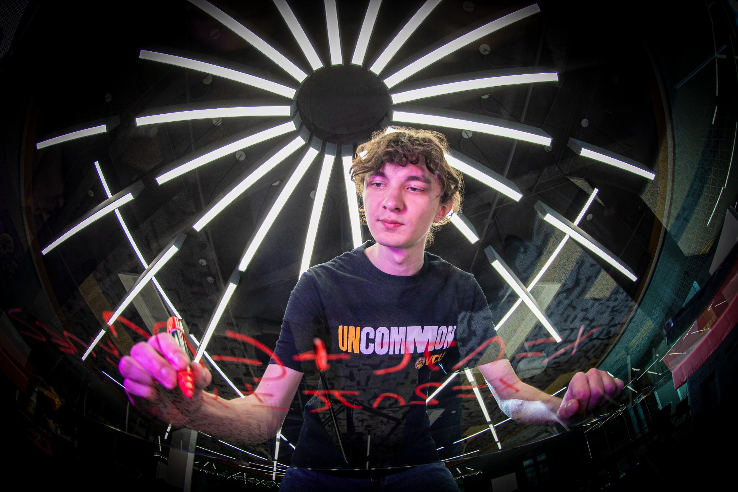 A photo fo a man wearing a shirt that says \"UNCOMMON\" writing an equation in red marker on a clear board.