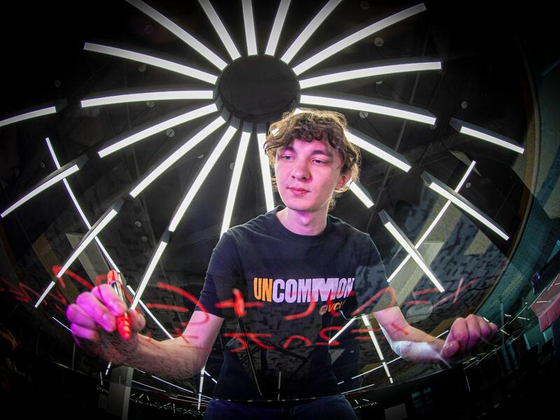 A photo fo a man wearing a shirt that says \"UNCOMMON\" writing an equation in red marker on a clear board.