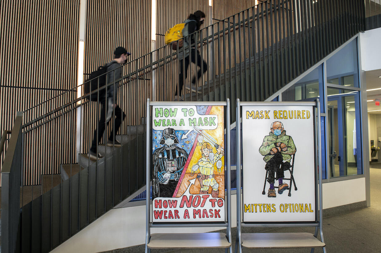 Students walk up the stairs at Cabell Library. A Star Wars-themed drawing on the left whiteboard states \"How to wear a mask. How to NOT wear a mask.\" A drawing on the right whiteboard of Sen. Bernie Sanders states \"Mask required, mittens optional.\"