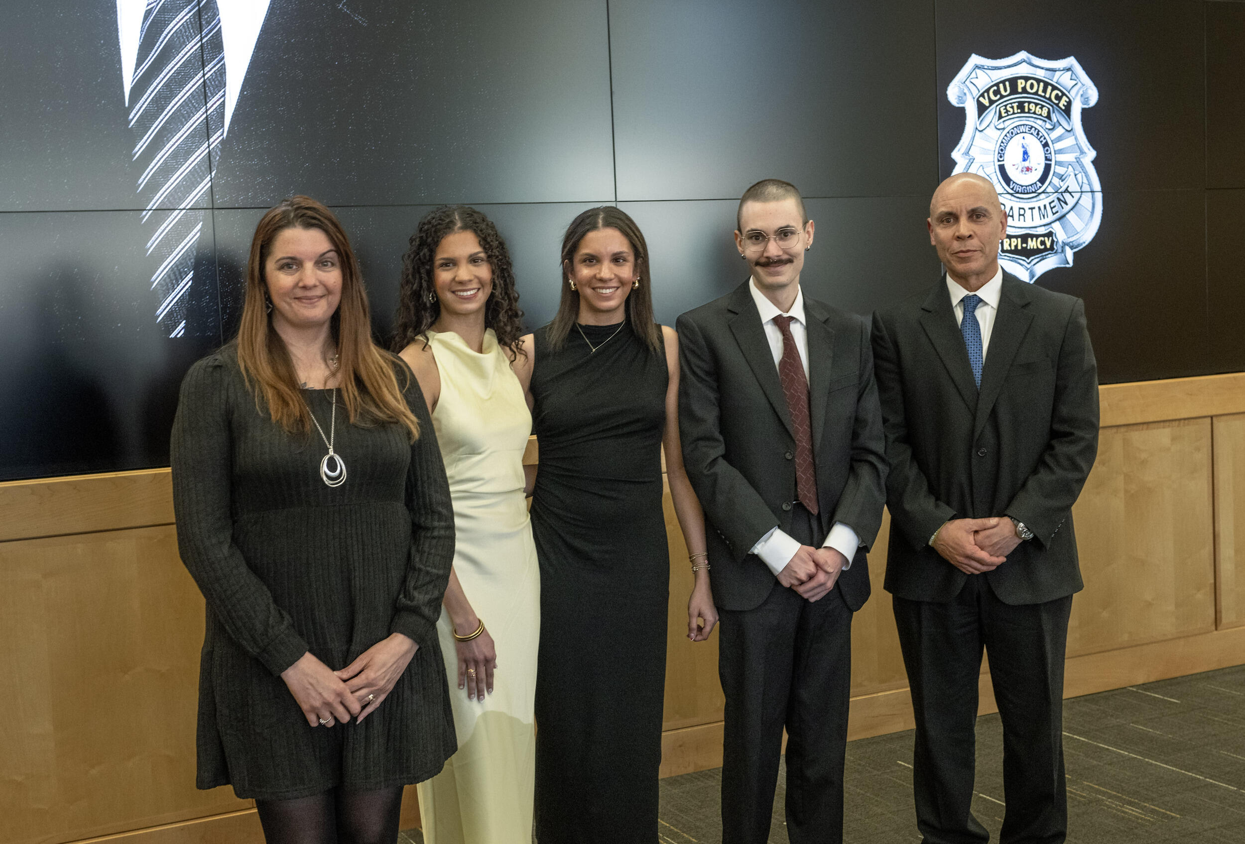 A group photo of three woman standing on the left and two men standing on the right. The women are wearing dresses and the men are wearing suit and ties. 