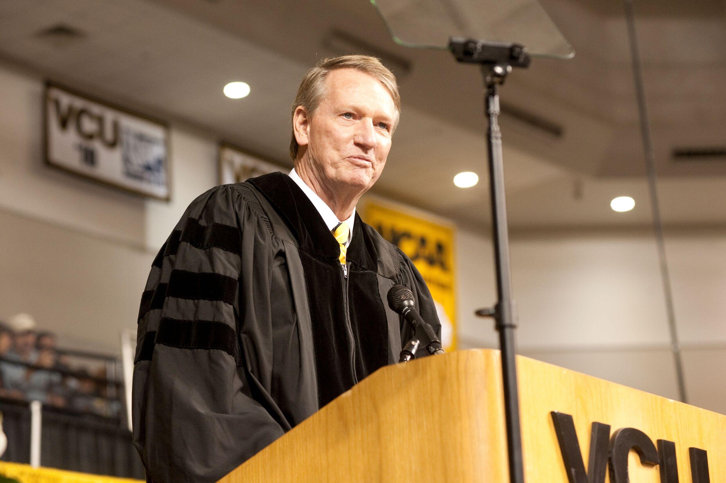 A man in a graduation gown speaks at a lectern.