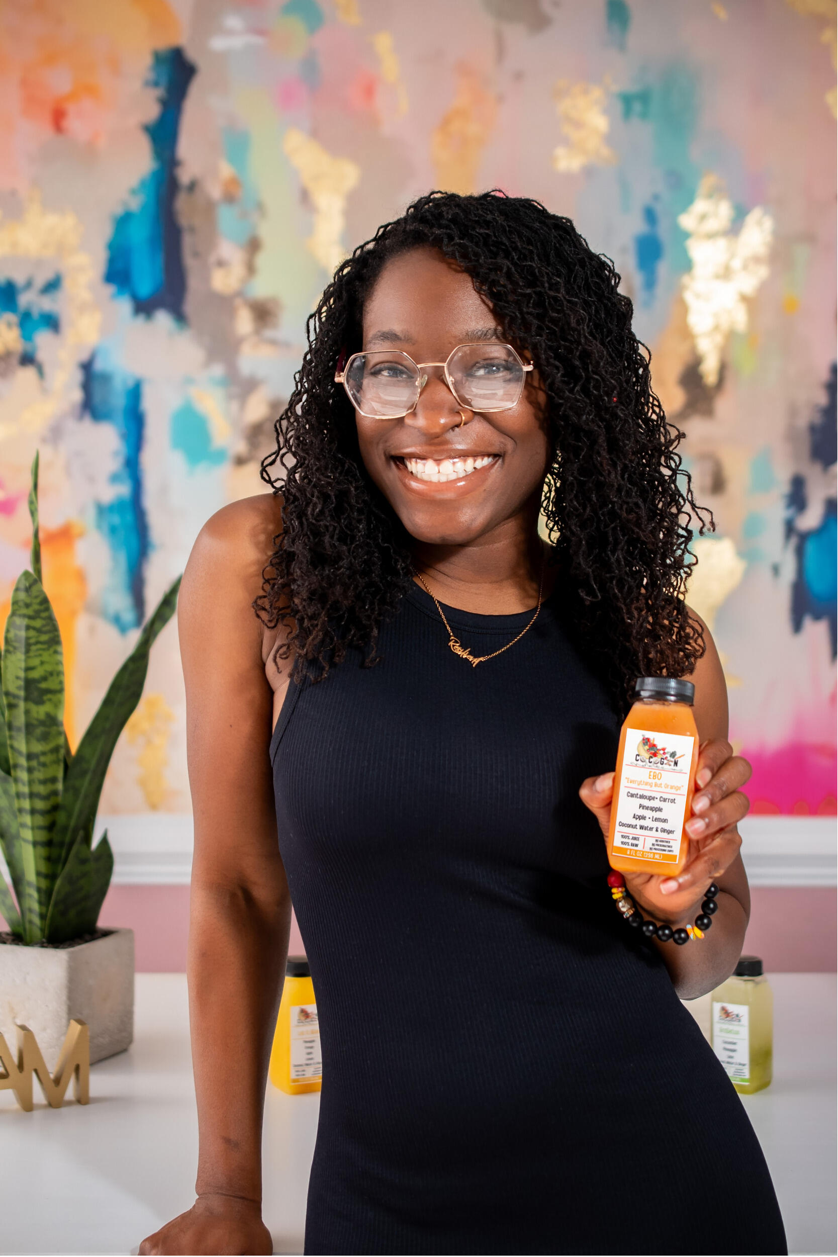 A photo of a woman from the waist up. She is smiling and holding a small bottle of orange juice in her right hand. 