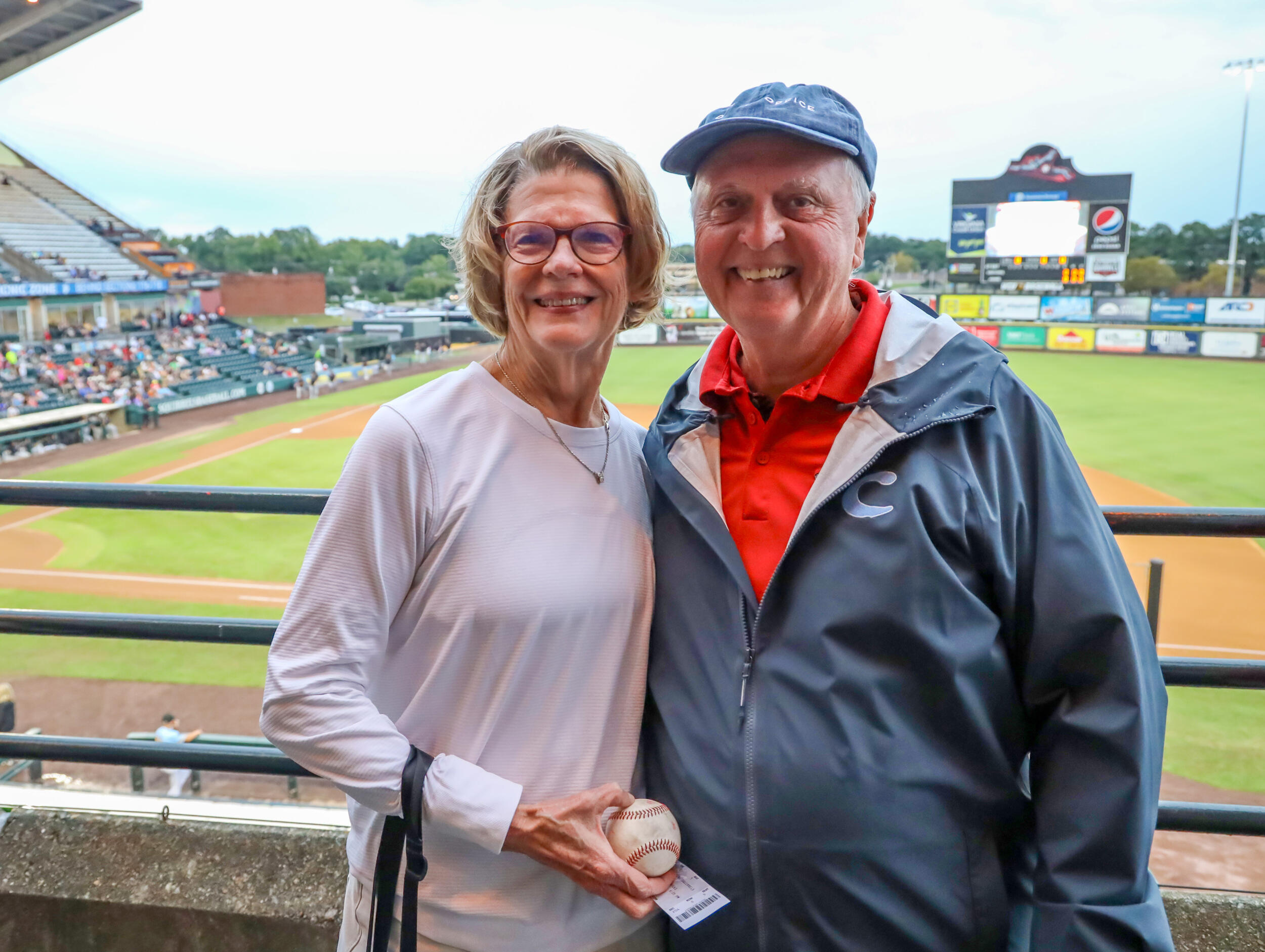 A photo of a man and a woman standing next to each other and smiling in front of a baseball field. The woman is holding a baseball in her left hand. 