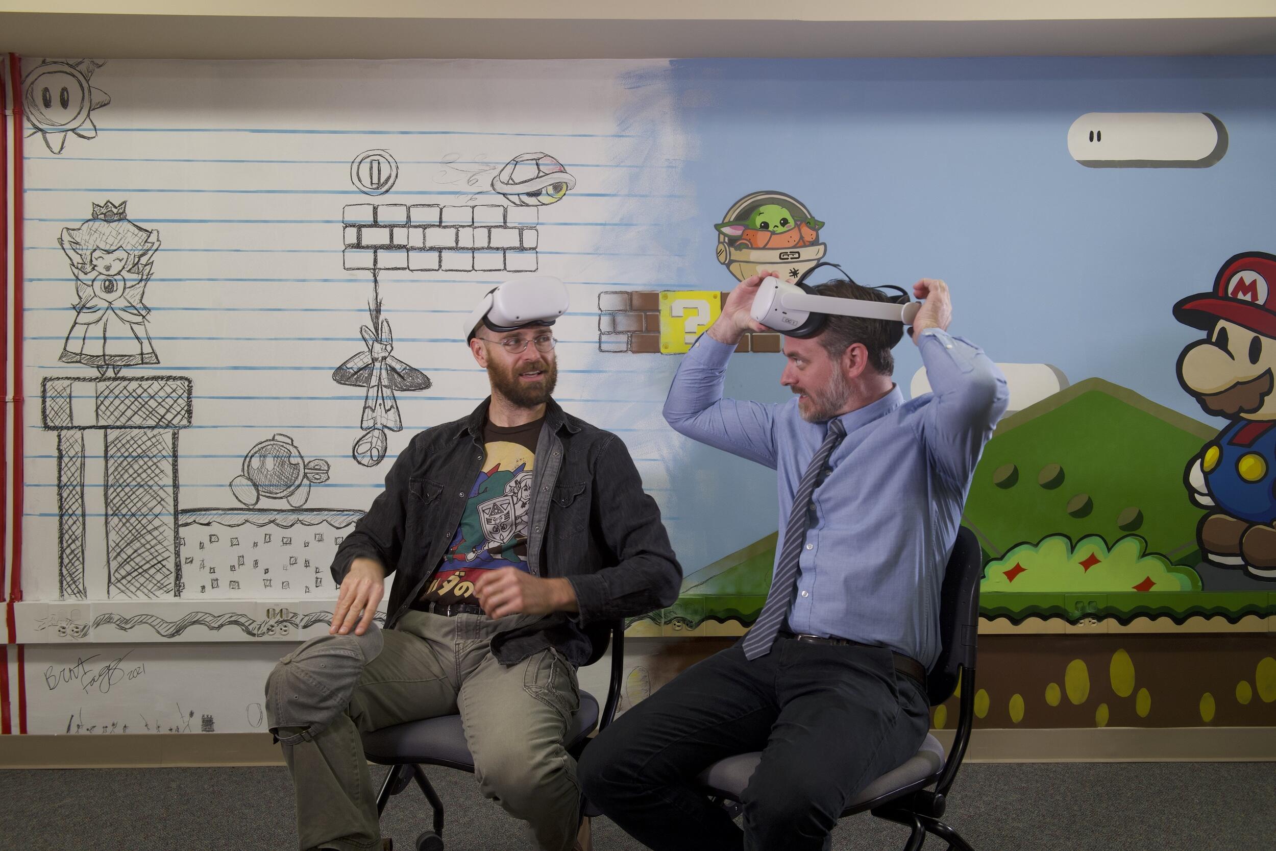 Two men sitting in chairs. The man on the left has a virtual reality goggles on his forehead, and the other man is placing them on 