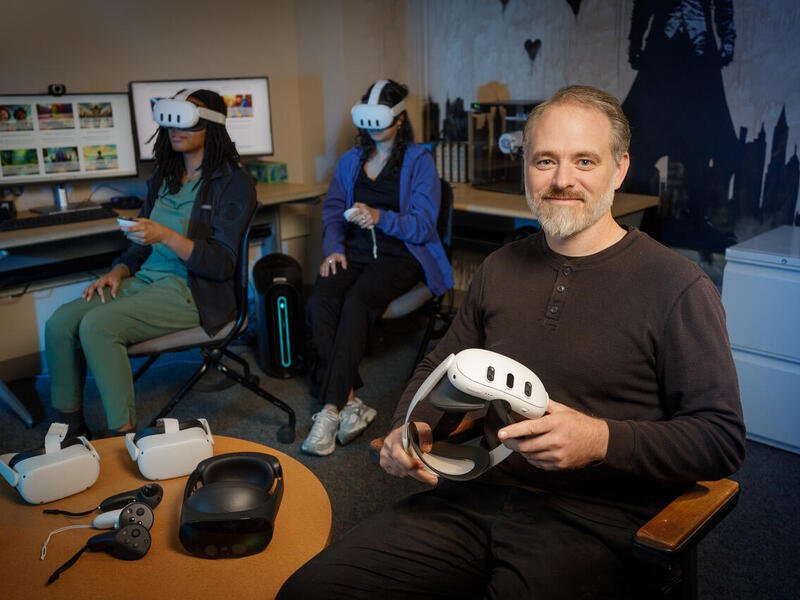 A photo of a man sitting in a chair holding a VR headset. Behind him are two women who are also sitting in chairs, but they are wearing VR headsets and holding VR controllers. On the table next to the man are three more VR headsets. 