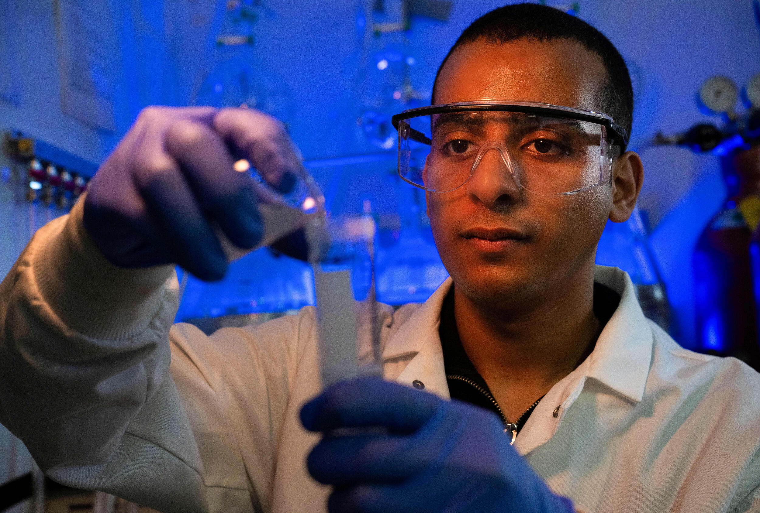 A man in safety goggles, gloves and a lab coat works in a lab.