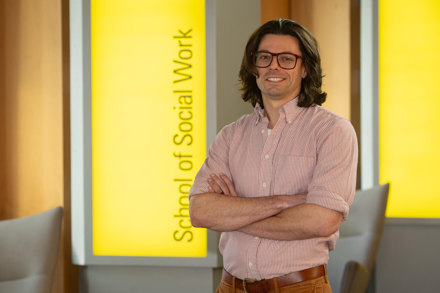 A photo of a man from the waist up. His arms are crossed against his chest. Behind the man is a yellow sign with black letters that says \"School of Social Work\"