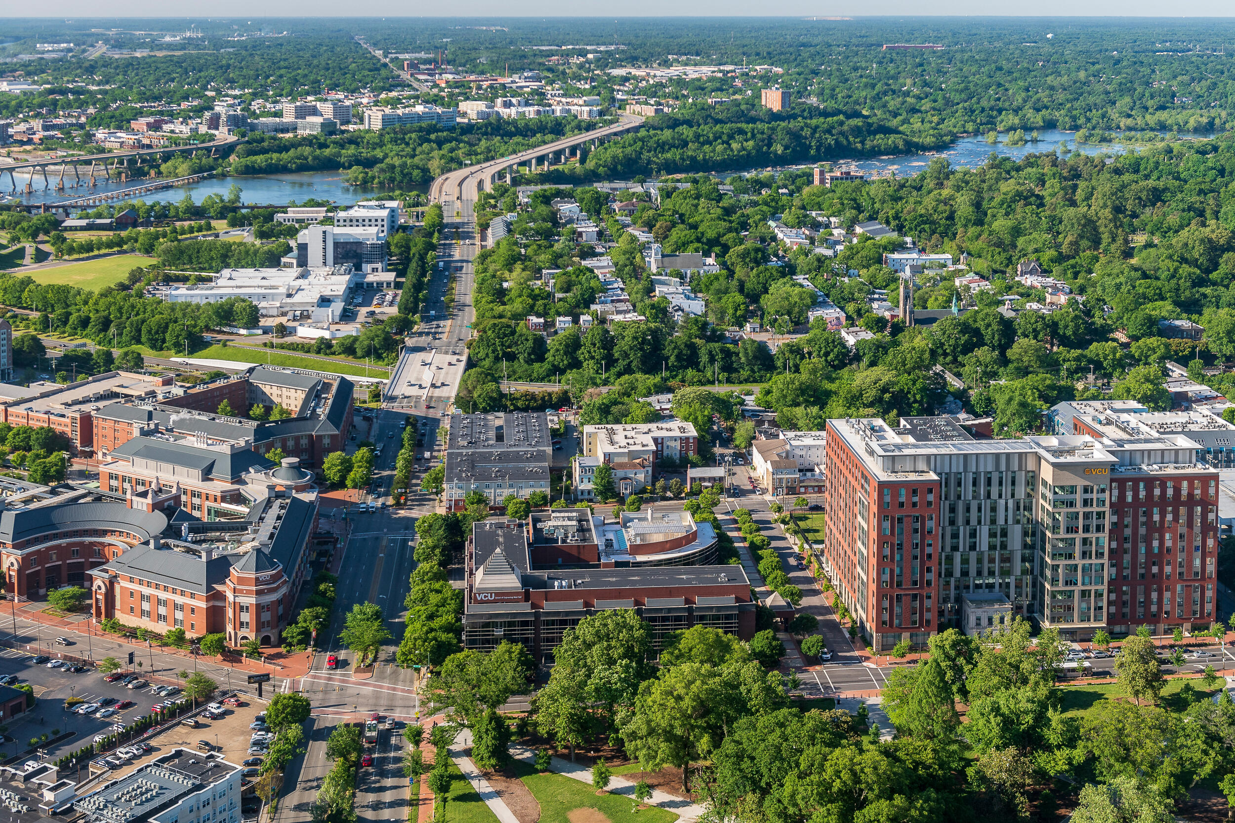 An aerial shot of VCU's Monroe Park campus facing south toward the James River. Several buildings on the southwest side of campus sit on either side of a large road that becomes a bridge across a river, with trees and buildings on either side of the road and of the river.