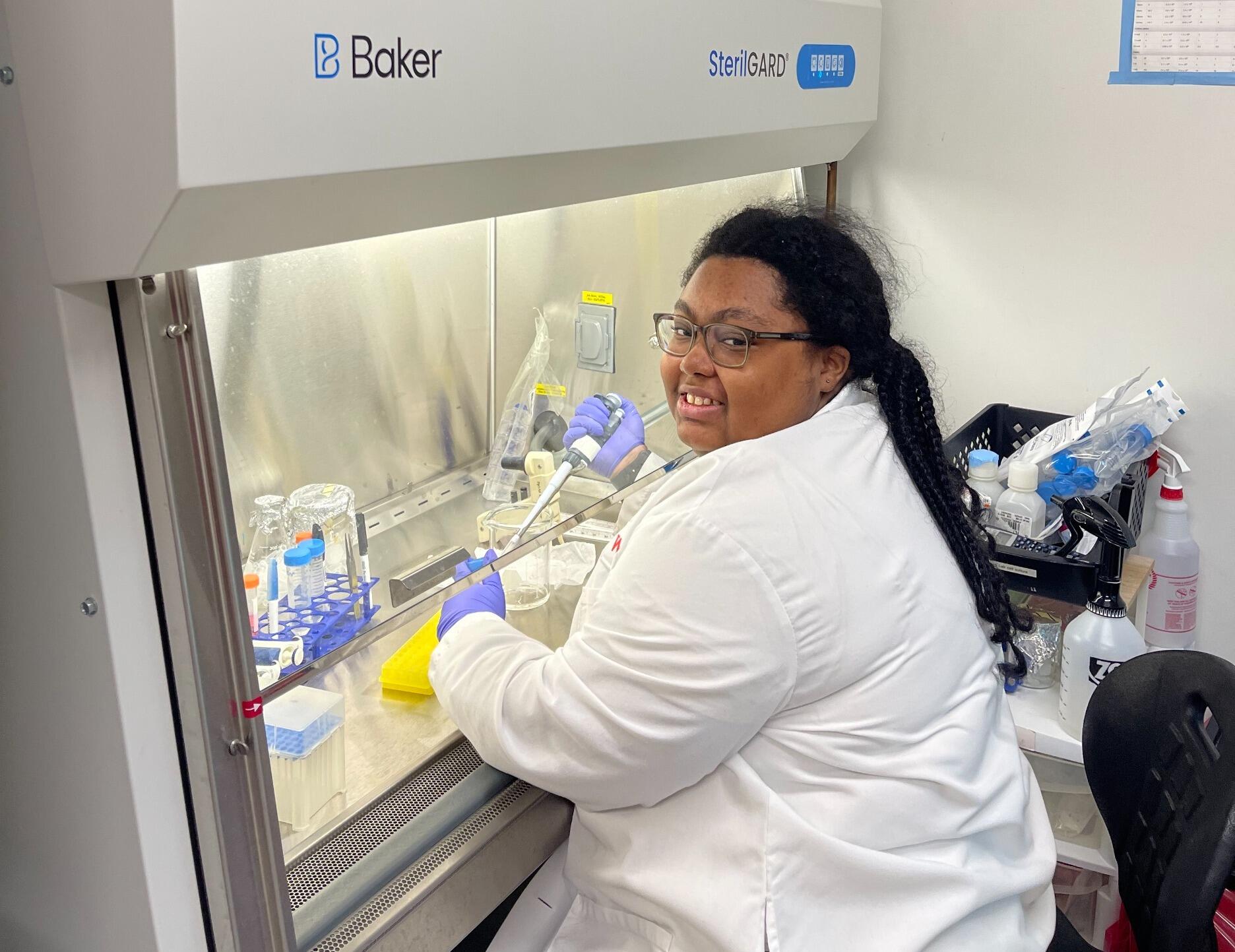 A photo of a woman sitting at a fume hood in a lab.