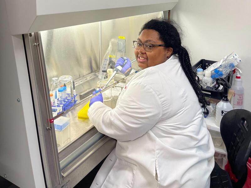 A photo of a woman sitting at a fume hood in a lab. 