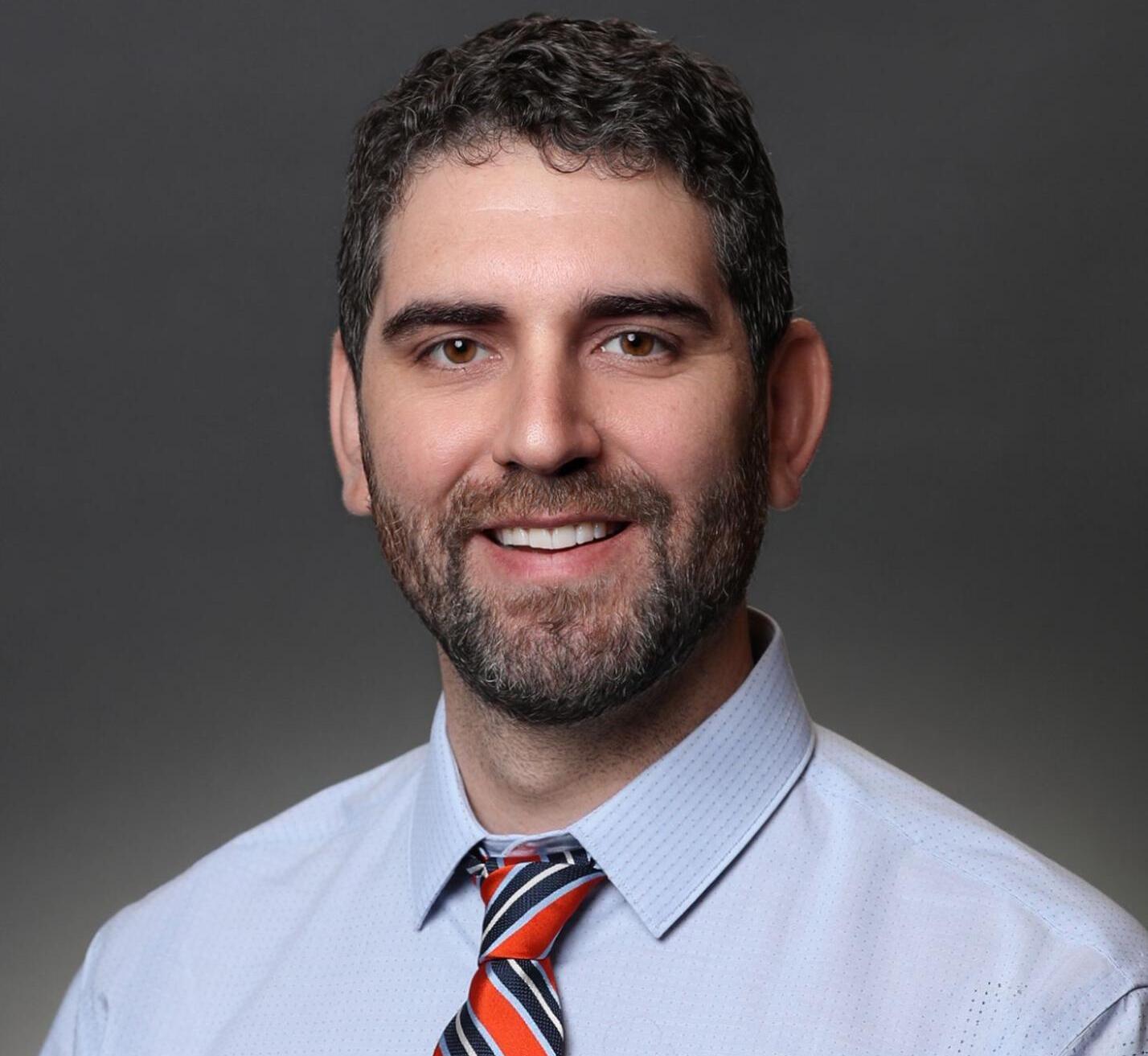 A photo of a man from the chest up. He is wearing a blue button up shirt and a red, blue, and white striped tie. 