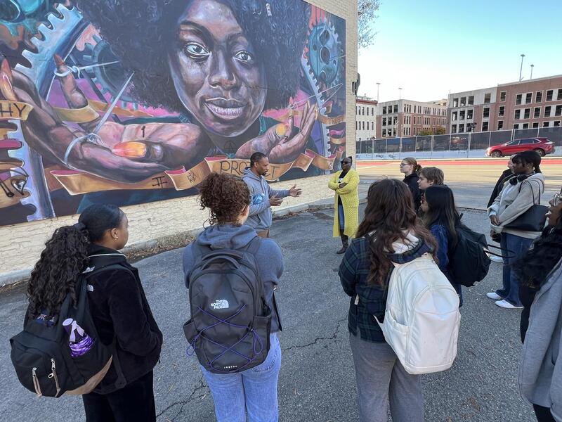 A photo of a man talking to a group of nine people who are in a semi-circle. Behind them is a mural of a woman. 