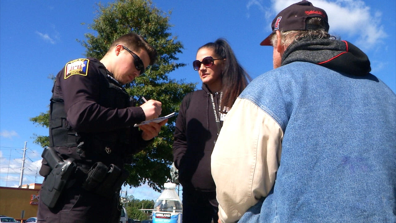 A police officer writes in a notebook while speaking with a woman in sunglasses. A man with his back to the camera in a baseball hat is also part of the conversation.