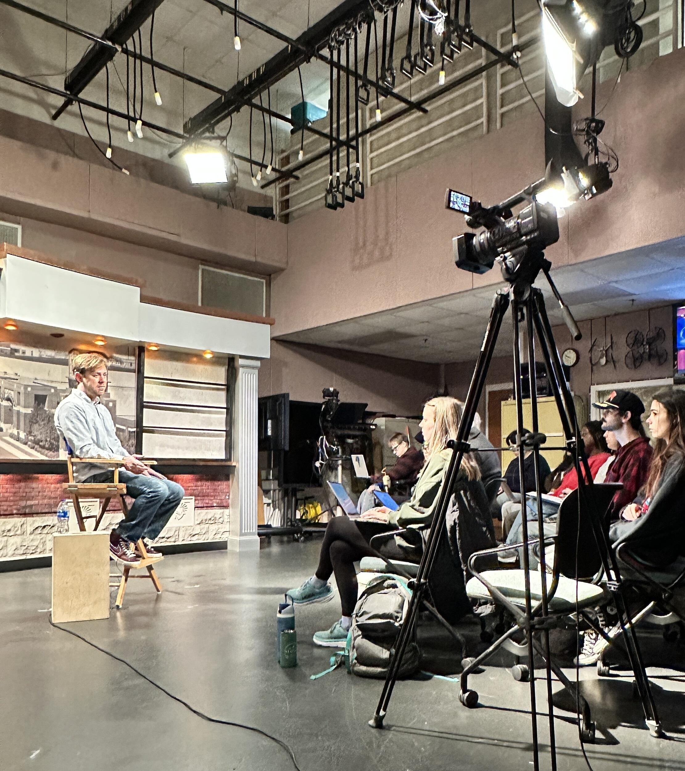 A photo of a man on a TV set. To the right is a camera and a woman sitting in a chair. 