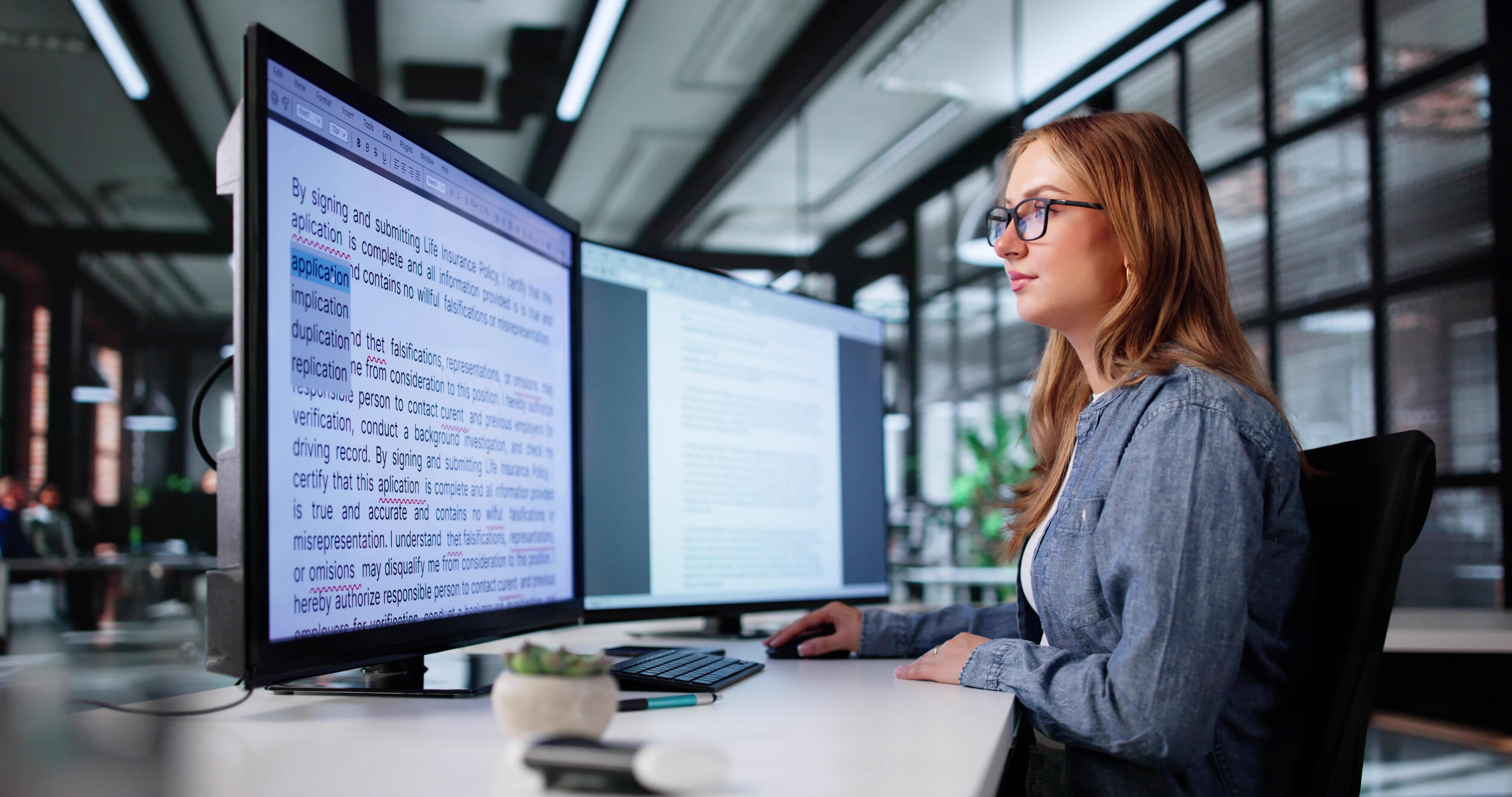A photo of a woman sitting at a desk with two computer monitors on it. She is looking at the monitor on the left which is covered in text.
