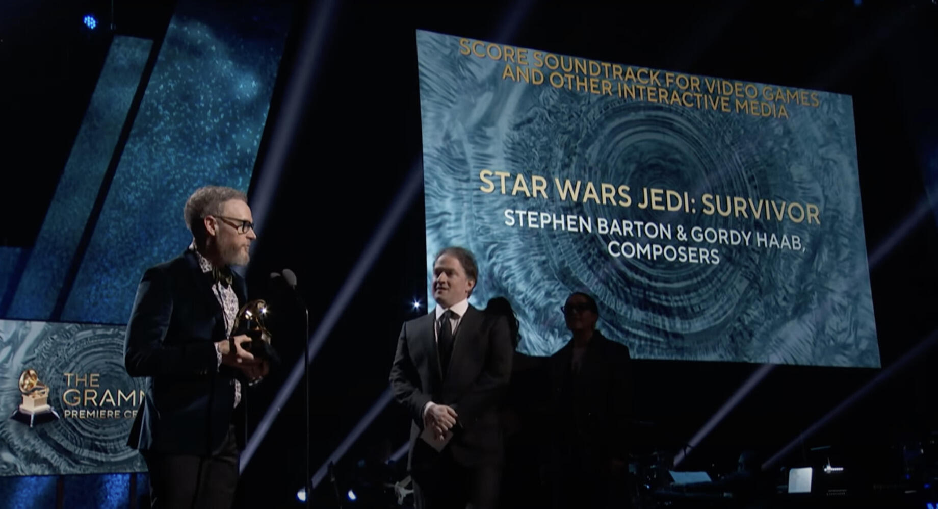 A photo of two men standin on a stage. The man on the left is standing holding an award while the man on the right watches him. Behind them is a screen that reads \"STAR WARS JEDI: SURVIVOR STEPHEN BARTON & GORDY HAAB COMPOSERS) 