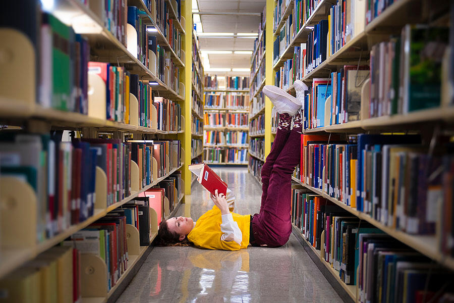 A photo of a woman laying in between two book shelves in a library. Her legs are pointed up and resting against one of the shelves. The woman is laying on her back and reading a book. 