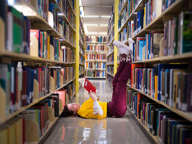 A photo of a woman laying in between two book shelves in a library. Her legs are pointed up and resting against one of the shelves. The woman is laying on her back and reading a book. 