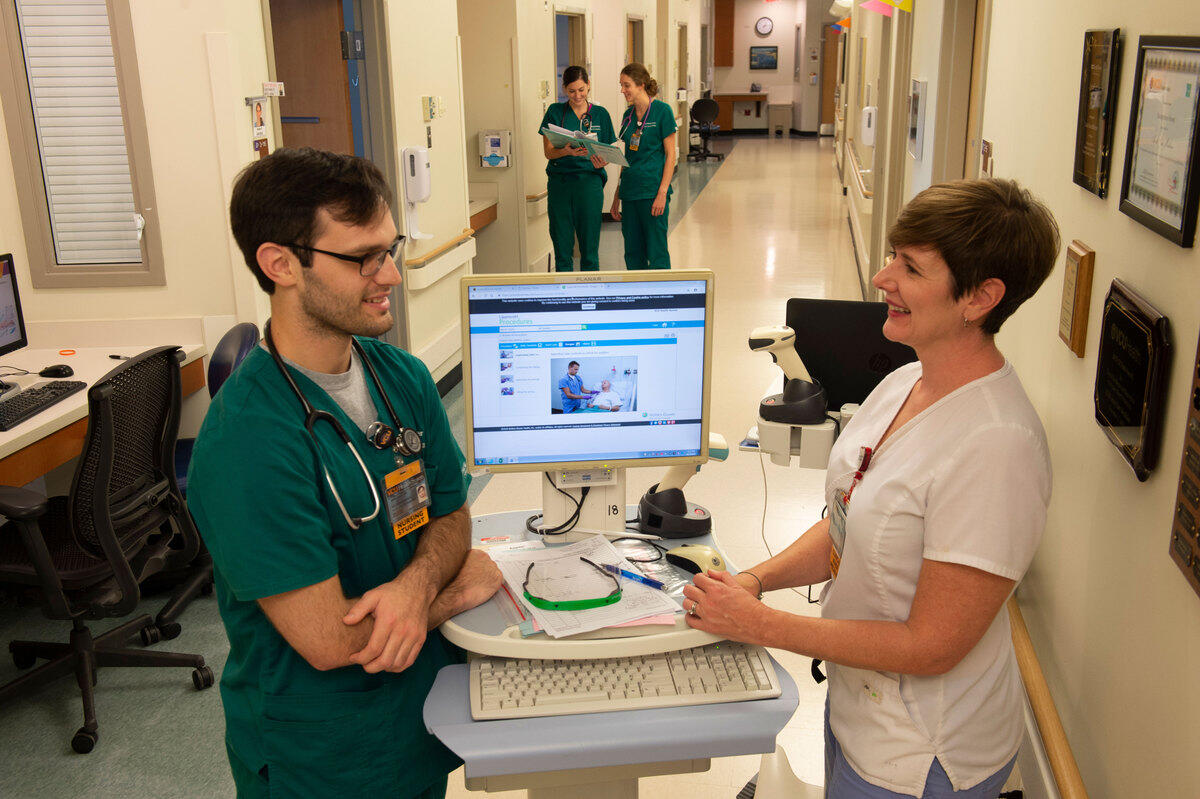 A woman (right) and a man (left) in nurse's clothes stand facing each other and speak in a health care facility hallway with a computer screen open between them.
