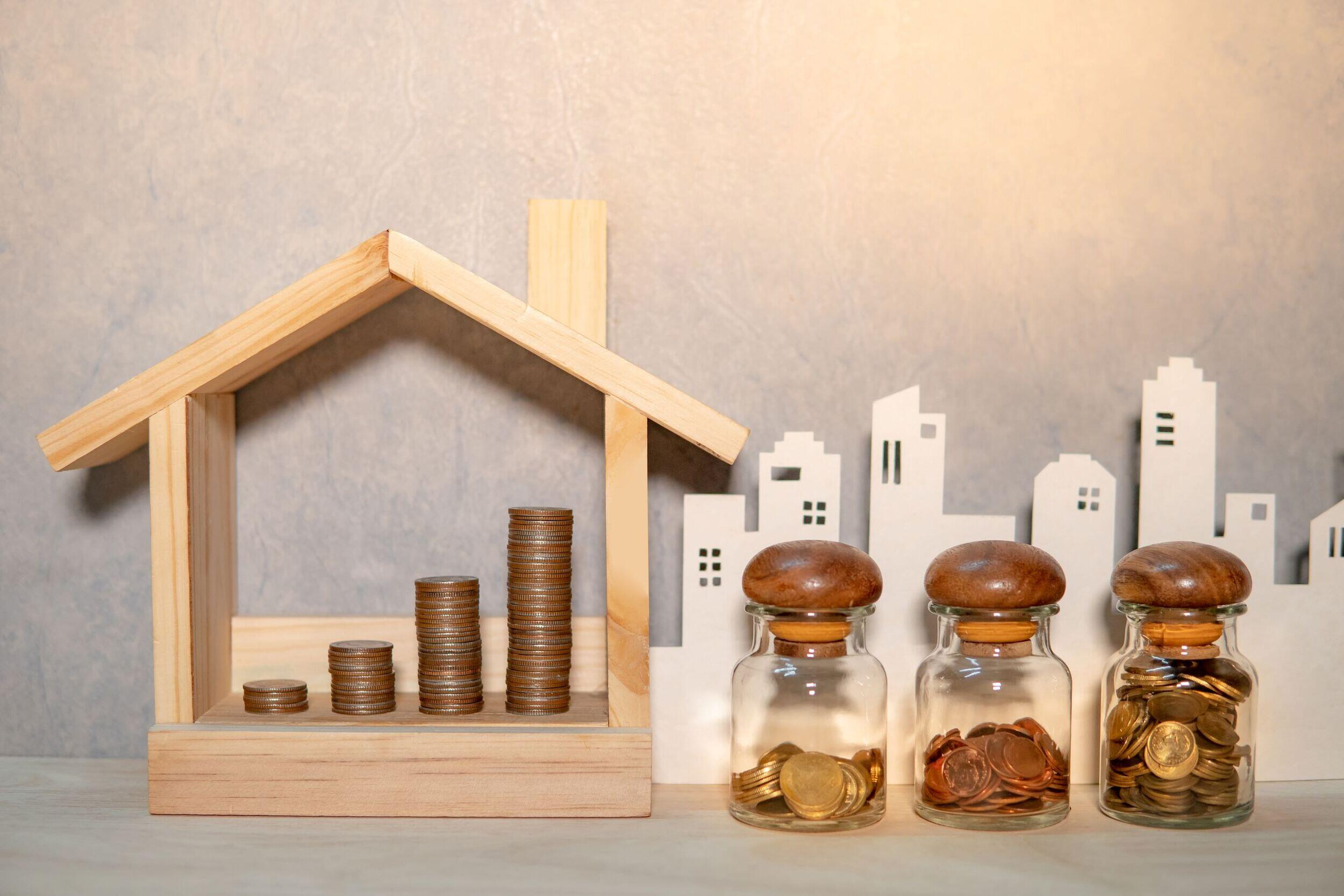 A small wooden house with stacks of coins inside. Next to it are three glass shakers with coins in them. Behind all that is the outlines of a city skyline.
