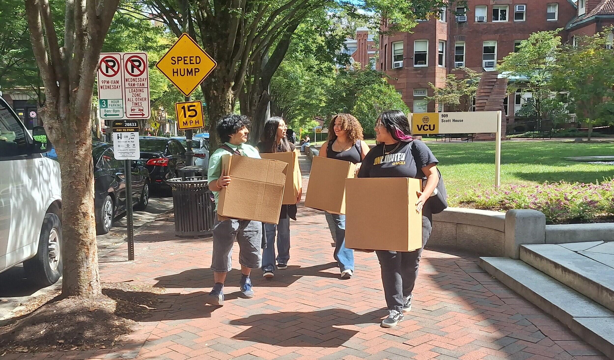 A photo of four students walking down a brick sidewalk carrying cardboard boxes. 