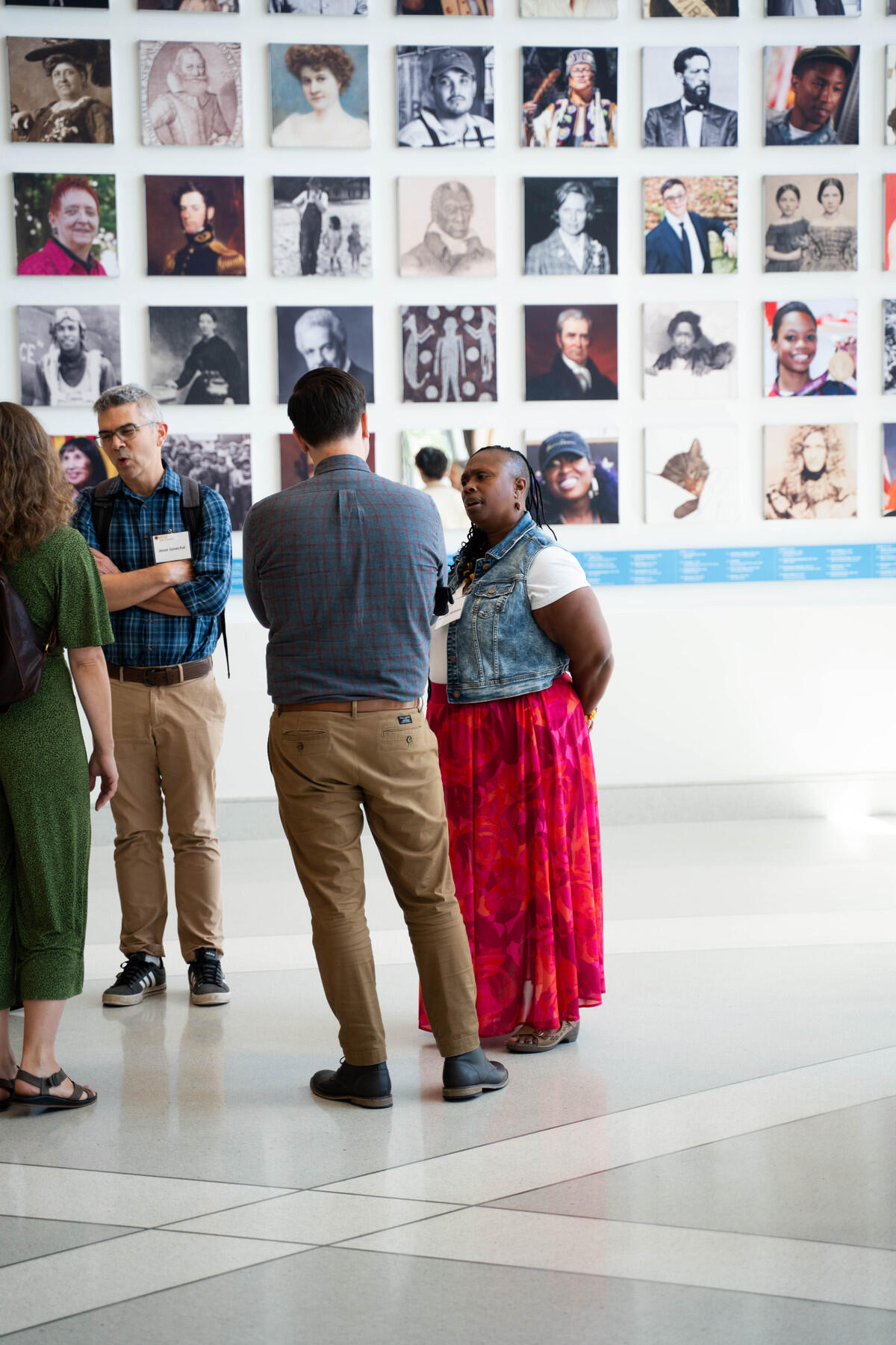 A photo of four people speaking in front of a wall covered in portaits of historic figures. 